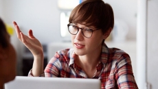 
  Person wearing glasses and a plaid shirt sitting at a computer with their hand up as though talking to someone on the computer
