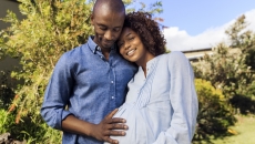 A pregnant person standing in a garden next to another person with their hand on the pregnant person's stomach