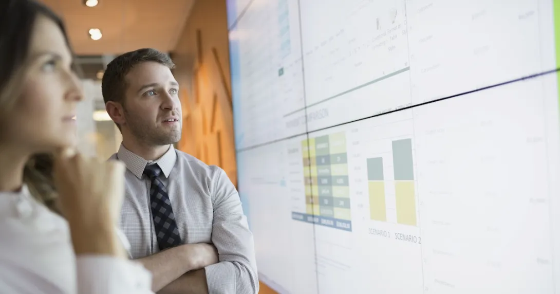 
  Two people standing up and looking at a large screen with graphs on it 
