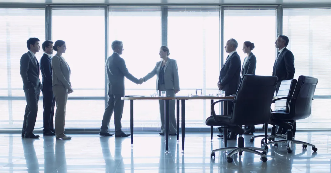Two people shaking hands in front of a big window with six other people watching