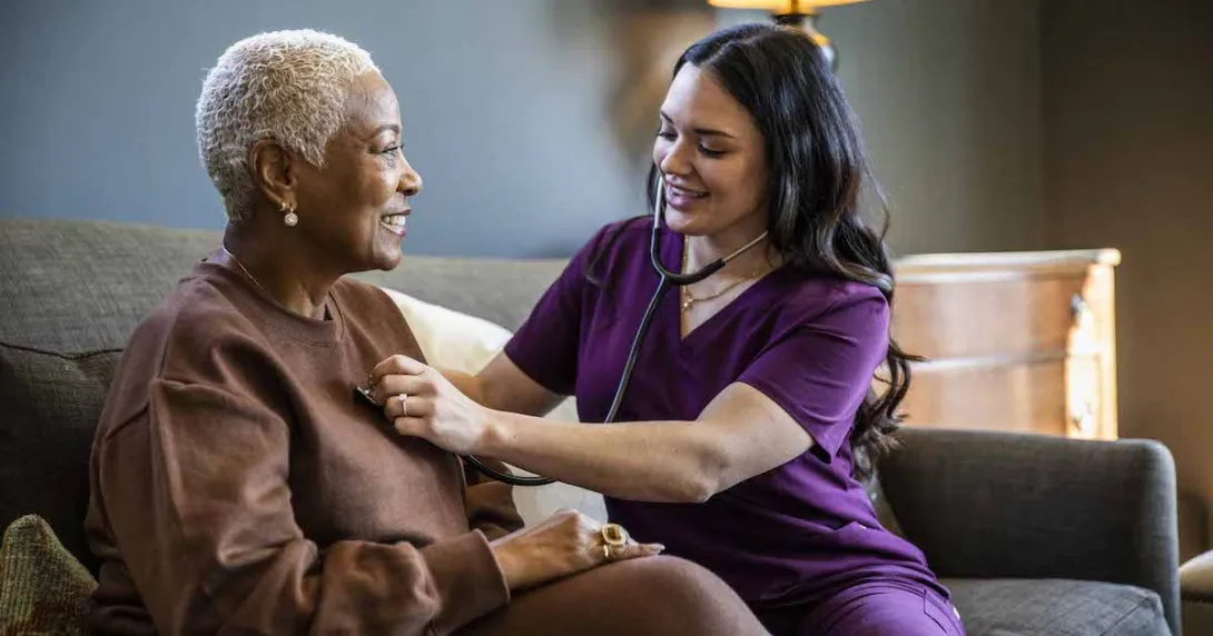 
  Healthcare provider sitting on a couch with a patient in their home checking their vitals. 

