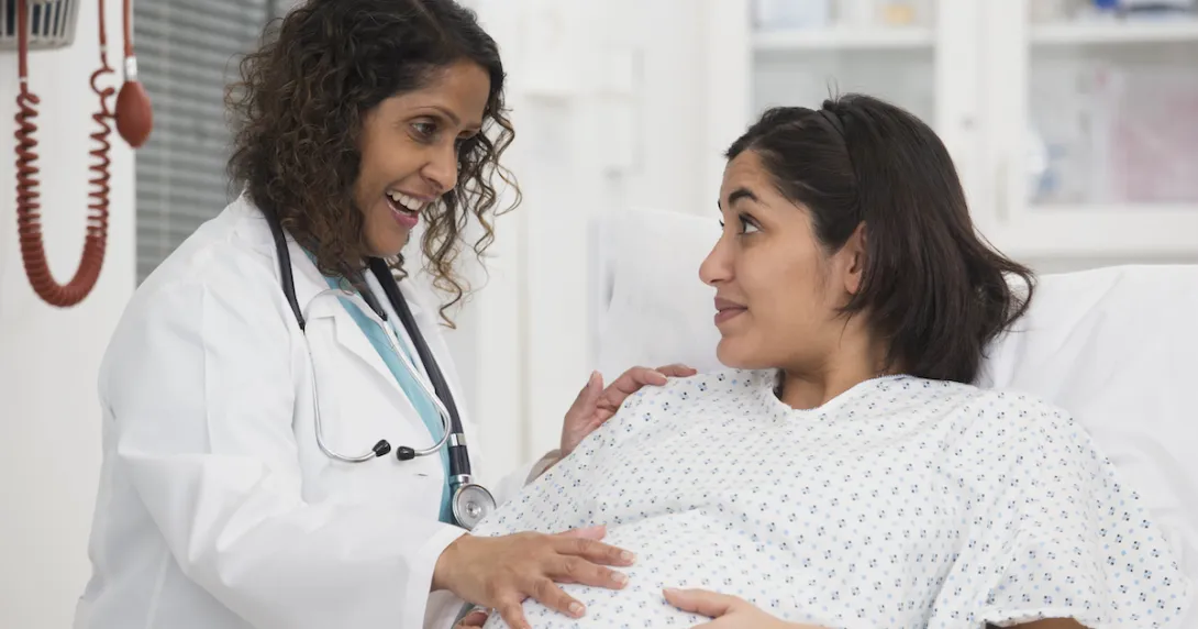 Healthcare worker standing beside a pregnant person lying in bed while both are touching the pregnant person's stomach
Healthcare worker standing beside a pregnant person lying in bed while both are touching the pregnant person's stomach