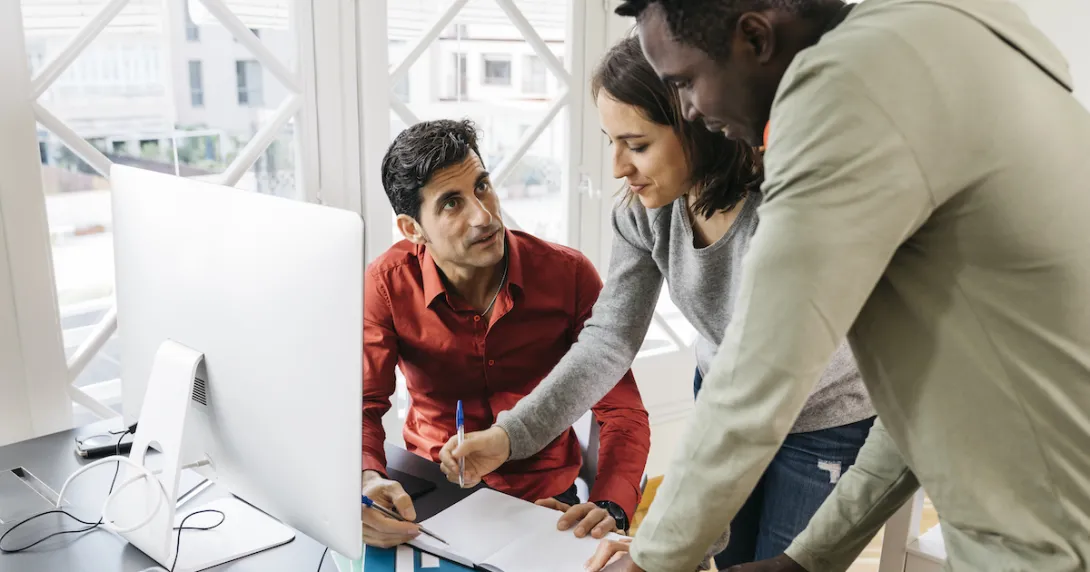 Person in a red shirt sitting at a desk in front of a computer with two people standing beside him looking at the computer
Person in a red shirt sitting at a desk in front of a computer with two people standing beside him looking at the computer