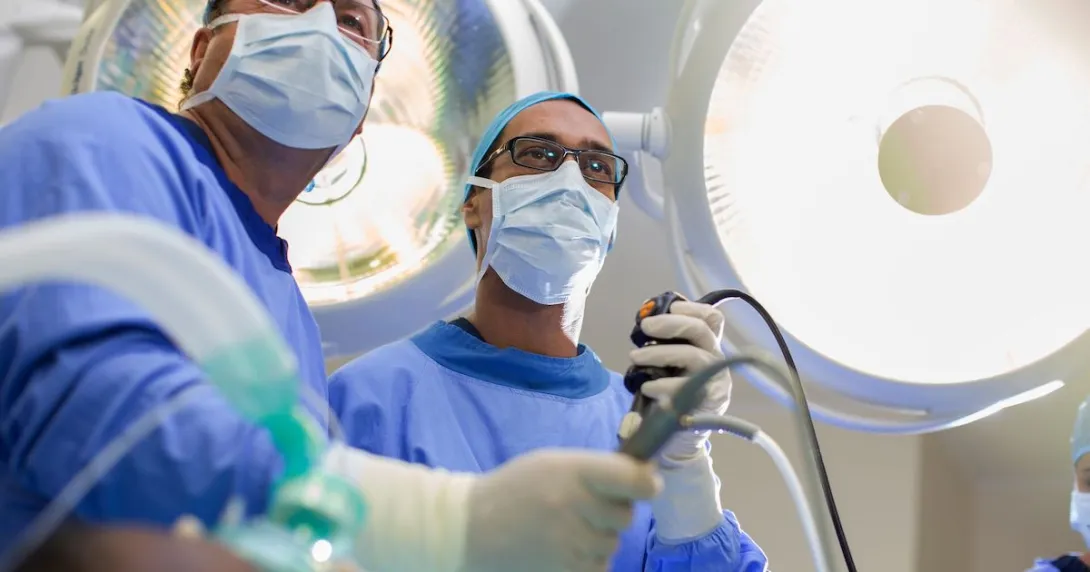 
  Two surgeons looking down at a camera with scrubs on while holding surgical tools
