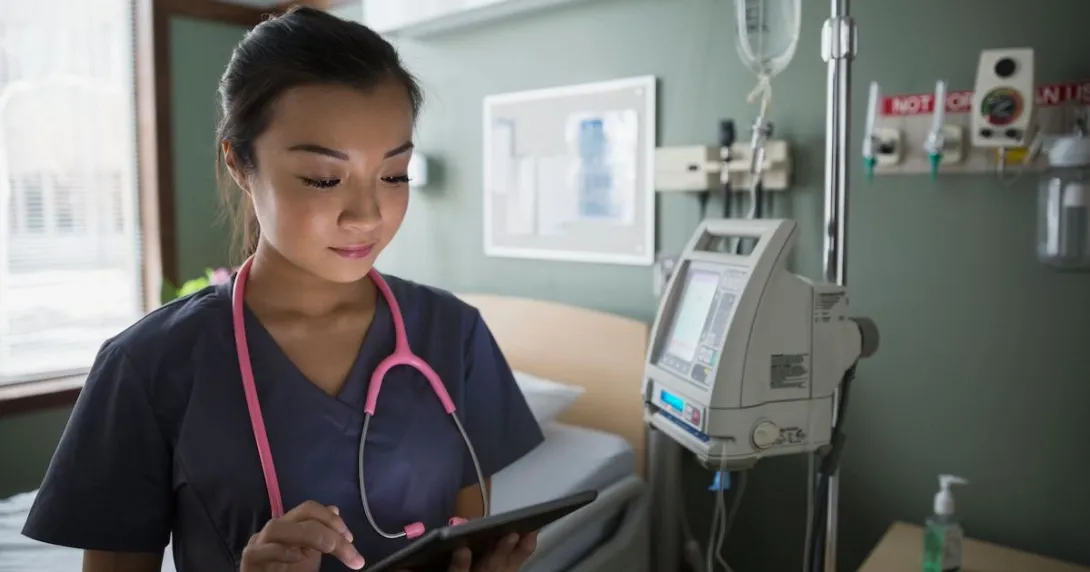 
  Healthcare provider standing in a hospital room wearing a pink stethoscope while holding a tablet 
