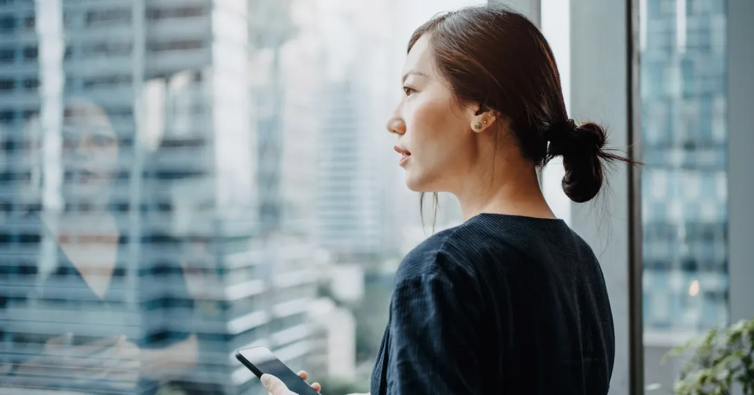 A person holding a smartphone looking out a window.